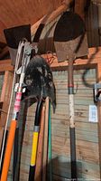 Photo showing a tree pruning saw with a curved serrated metal blade and an orange handle, along with two shovels/spades with metal heads and wooden or fiberglass handles hanging on a wall.