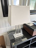 Full view of the lamp showing white rectangular fabric shade and clear crystal base with silver trim, lamp positioned on a black shelf.