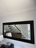 Full view of the large rectangular mirror reflecting stairway and wall behind it, showing black framed bevelled edge glass.