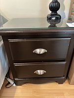 Front view of the dark wood night stand showing two drawers with silver cup pulls and glass top.