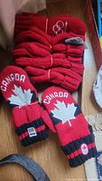 Six pairs of red Canada Olympic mittens with maple leaf and Canada embroidery, several pairs tied with rubber bands, placed on wooden floor showing cuffs with Olympic and Canada logos.
