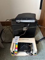 Two Polaroid Land Cameras shown together. One is boxed OneStep model in original rainbow stripe box, the other a vintage Land Camera with black textured body and leather strap placed on top of a carrying case.