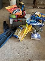 Photo of blue swim flippers, yellow scuba mask with snorkel, box for floating lounger, floating pizza slice box, assorted pool toys in gray plastic bin
