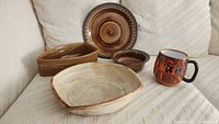 Four pieces of hand crafted pottery including plate, matching bowl, boat-shaped bowl, large squared bowl, and a decorated ceramic mug placed on a white fabric surface.