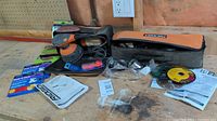 Photo of Rigid variable speed random orbit sander, angle grinder, sanding discs, grinding blades, ear plugs, and operator manuals laid out on a wooden surface.