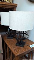 Pair of table lamps with linen drum shades on dark metal orbital bases, shown together on a wooden table.