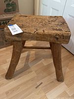 Front-side angle view of handmade wooden stool showing thick seat and three sturdy legs, rough wood texture, natural cracks and holes