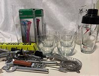Barware lot contents including 6 clear glasses, cocktail shaker with black lid and starburst design, clear pitcher, various cocktail tools with wooden handles, box of colorful stirring rods.