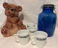 Photo showing Gilner bear cookie jar, four white milk glass mugs with blue floral band, and large cobalt blue glass cookie jar.