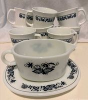 Front view showing 8 white mugs with blue floral band and the gravy boat with saucer with blue floral design on white glass.