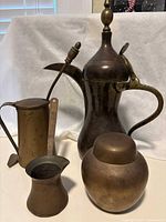 Four brass items against white backdrop: antique smudge pot with fire starter stick, Turkish coffee pot, brass urn, smaller brass coffee pot, showing surface wear and tarnishing