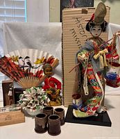 Full view of Japanese Geisha figurine with musician figurine, ceramic flower arrangement, brown sake cups, decorative fan, wooden toothpick box, and souvenir box.