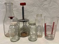 Overall view of six vintage glass measuring and storage containers including Universal Store bottle, measuring cup with metal lid, baby bottle, small jars, and measuring glass.