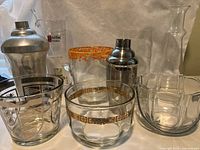 Overview photo showing all items in the barware lot including cocktail shakers, mixing glasses, decorative glassware with gold and orange accents, and cut-glass decanter.