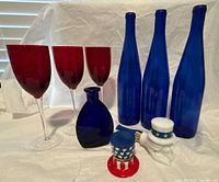Red wine glasses, blue glass bottles and vase, ceramic Uncle Sam pipe and trinket box on white cloth background