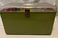 Closed avocado green plastic sewing box with clear lid and metal clasp closure, showing lid cracks.