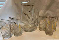 Full set photo showing pitcher with six matching glasses, all clear glass with gold and white leaf pattern and gold rims.
