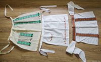 Three aprons laid out flat on wood floor, showing the green wool apron, white embroidered apron, and white apron with woven belts.