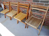 Four wooden folding chairs lined up against a brick wall, showing the side and front profiles. Chairs have wooden slatted seats and backs with visible edge wear.