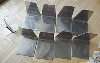 Nine metal bookends arranged on a wooden floor, showing each bookend's shape including flat base and angled vertical support. Surface reflections and slight wear visible.
