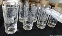 Group of eight clear etched floral pattern glasses, 7.5 inches tall, arranged in rows on black surface.