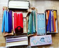 Overview of the assortment of colorful taper candles laid out on wood floor in various packaging and loose, including colors red, blue, green, purple, orange, white, and cream. Some are spiral shaped and some straight.