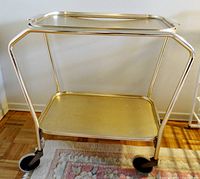 Full view of the pale gold metal tea trolley showing its frame, two shelves, and four wheels.