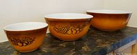 Three Pyrex Old Orchard mixing bowls in varying sizes shown side by side, showcasing vintage fruit pattern on amber gradient glass exterior.