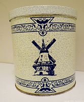 Front view of vintage cookie tin decorated with blue Dutch windmill and ornate borders on speckled beige metal surface.