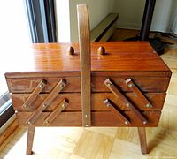 Front view of vintage wooden sewing box with handle and legs, showing expanding side compartments closed.