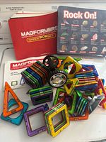 Overview of multicolor magformers magnetic tiles including squares, triangles, and accessory wheels spread out with instruction manuals and storage pouch visible.