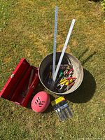 Photo showing red metal toolbox, pink-red XS/S helmet, metal bucket with assorted hand tools, and ProPainter paintbrush set on grass.