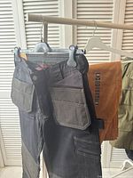 Black and brown Tough Duck work pants hanging on a rack showing the front with multiple utility pockets and sturdy stitching.