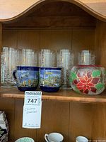 Shelf display showing five clear etched holly drinking glasses, two blue Christmas-themed coffee mugs from Hamburg Christmas Mart, and a large round poinsettia candle holder.