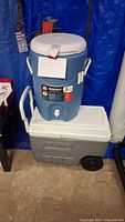 Two coolers stacked; top is blue Igloo 5 gallon beverage cooler with spigot, bottom is light gray Coleman wheeled cooler.
