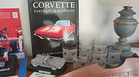 Photo showing the book, clear glass model car, and six Corvette-themed glasses on a wooden surface next to a silver lamp.
