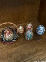Overview of the five decorative eggs arranged on a wooden surface with one egg placed inside a small wicker basket filled with shredded paper filler.