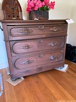 Front view of wooden dresser showing three drawers with detailed wooden pulls and decorative recessed panels on drawer fronts. Markings visible on top surface. Set on wooden roller wheels.