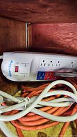 Photo of white surge protector power strip with multiple outlets, red power switch, and orange extension cords piled around on wood shelf