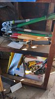 Photo showing green-handled pruning loppers, hand pruners with wooden handles, and various garden hand tools on a wooden shelf.