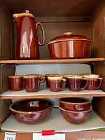 Shelf display of Hull pottery items including tall coffee pot with lid, covered casserole, two bowls with chips, and eight brown McCoy coffee mugs.