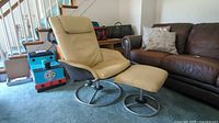 Beige Ikea leather look lounger chair with metal swivel base and matching ottoman in a carpeted room, next to a brown leather sofa and children's toy box.
