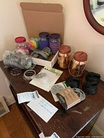 Overview of Mason jars with specialty lids and various accessories arranged on a dark wood table near a mirror. Includes jars with colored lids, copper lantern lids, wire lid holders, and silicone grippers.
