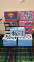 Photo showing stacked metal pencil boxes: one Superman design on top, five black and silver boxes stacked, two floral chevron boxes, and five blue penguin boxes.