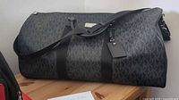 Front side of black Michael Kors duffle bag showing the MK logo pattern and removable shoulder strap.