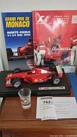 Photo showing the F2001 model Ferrari car with decals and the two Grand Prix de Monaco programs behind it, plus the shot glass in front. Includes a lot note with details.