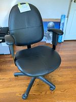Front right angled view of the black leather-like office chair showing the seat, backrest, adjustable height lever, and armrests on wooden floor.