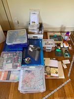 Overview of lot items on wooden floor including trimmer box, storage boxes with washi tape, cardstock pack, adhesives, punches, magnetic die storage case, assorted paper packs in cardboard box