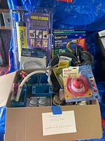 Photo of assorted plumbing and electrical supplies including power plunger, gas installation kit, drain cleaner, valves, and blue electrical junction boxes in a box