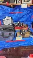 Overview of lot showing power tools, drill bits, accessories, fabric tool bags, and small metal toolbox on blue tarp.